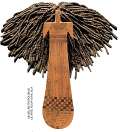 Imagem: Fotografia. Uma boneca feita de madeira com corpo alongado e fino, curva na extremidade e geométrico na região da cabeça, com cabelo trançado e volumoso. Na superfície do corpo há desenhos entalhados.  Fim da imagem.