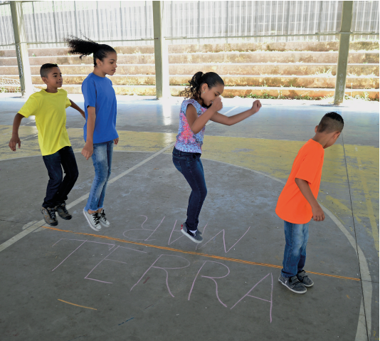 Imagem: Fotografia. Em uma quadra coberta, quatro crianças brincam pulando enfileirados um atrás do outros. No chão, há uma linha que divide de um lado uma parte na qual está escrito MAR e outra na qual está escrito TERRA.  Fim da imagem.