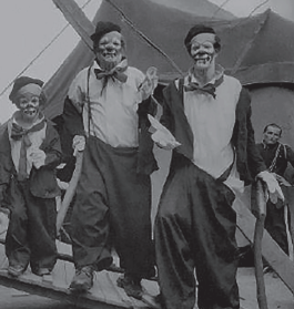 Imagem: Fotografia em preto e branco. Três palhaços lado a lado. Os rostos estão pintados, eles usam pequenos chapéus, gravatas compridas no pescoço e roupas largas.  Fim da imagem.