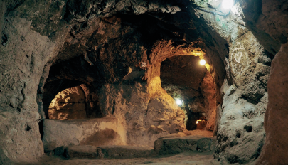 Imagem: Fotografia. Interior de uma caverna com chão batido, passagens arqueadas escavadas e pouca iluminação.  Fim da imagem.