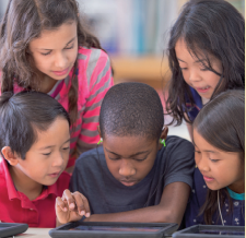 Imagem: Fotografia. Cinco crianças estão entretidas observando a tela de um dispositivo sobre uma mesa.  Fim da imagem.
