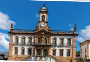 Imagem: Fotografia. Fachada de um prédio antigo com acesso por duas escadas laterais, dois andares com janelas retangulares, entrada com portas arqueadas e sacadas. Em uma delas há a bandeira do Brasil hasteada.  Há um terraço com parapeito, esculturas nas laterais e uma torre ao centro onde há um sino e um relógio analógico.  Fim da imagem.