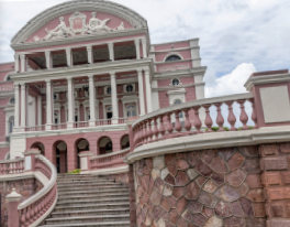 Imagem: Fotografia. Fachada de um prédio com entrada que apresenta um lance de escadas em formato curvo e prédio com três andares com arcos e muitas colunas. A parede do lado de fora apresenta tijolos e pedras aparentes Fim da imagem.