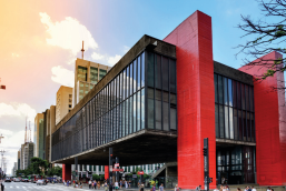 Imagem: Fotografia. Fachada de um prédio suspenso por colunas com formato retangular e estrutura de vidro. As colunas são grossas, vermelhas e são duas em cada lateral.  Fim da imagem.