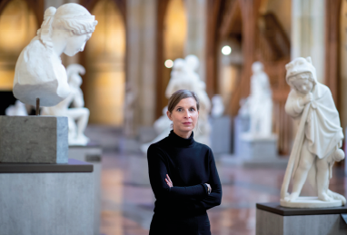 Imagem: Fotografia. Uma mulher loira de cabelo liso e pele clara veste preto e está com os braços cruzados ao centro de um amplo espaço com muitas esculturas brancas expostas ao redor. Fim da imagem.