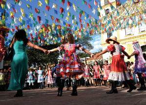 Imagem: Fotografia. Na rua, durante o dia, um grupo de pessoas dança em roda. As meninas usam bota e vestido longo colorido, algumas usam chapéu e outras estão com cabelo preso. No espaço, há muitas bandeirolas coloridas penduradas. Fim da imagem.