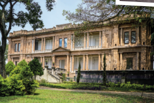 Imagem: Fotografia. Fachada de um prédio com dois andares que apresenta muitas janelas e portas com formato retangular, entrada com pequeno lance com escadas e colunas ao longo da estrutura. À frente do prédio, há um jardim com área verde.  Fim da imagem.