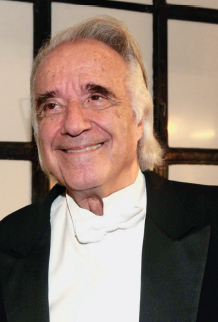 Imagem: Fotografia. Retrato de um homem de pele clara e cabelo grisalho liso e comprido. Ele sorri e veste roupa formal.  Fim da imagem.