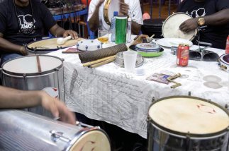 Imagem: Fotografia. Uma mesa onde estão alguns instrumentos, como tamborim e agogô, e ao redor da qual algumas pessoas tocam cuica, pandeiro e surdo. Fim da imagem.