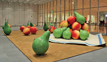Imagem: Fotografia. Em uma ampla sala com pé direito alto, bem iluminado e com parede de vidro há uma composição com nectarinas e peras gigantes.  Fim da imagem.