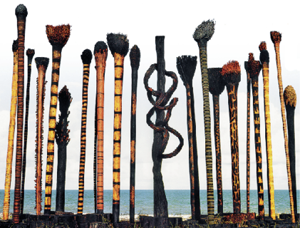 Imagem: Fotografia. Em primeiro plano, sobre pequenas toras pretas, uma composição com cipós, raízes e caules com superfície colorida, com destaque para o amarelo, laranja, verde e preto. Eles estão lado a lado fixados sobre as toras. Ao centro, destaque para um tronco escuro mais grosso envolto por uma raiz retorcida. Ao fundo, o mar.  Fim da imagem.