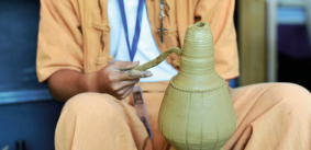 Imagem: Fotografia. Destaque de uma pessoa que usa um instrumento metálico pequeno na superfície de um objeto esculpido em argila. Fim da imagem.