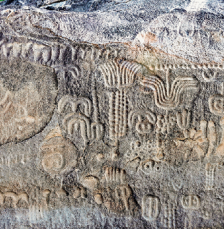 Imagem: Fotografia. Destaque da superfície de uma pedra grande e de aparência espessa sobre a qual há inscrições com símbolos diversos. Fim da imagem.