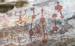 Imagem: Fotografia. Destaque de uma parede rochosa que apresenta desenhos de animais ao lado de figuras humanas ao redor de uma fogueira. A cor predominante é marrom.  Fim da imagem.