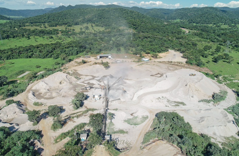 Imagem: Fotografia. Vista do alto de local com solo de cor bege-claro. Ao redor, local com grama e vegetação de cor verde. Na parte inferior, construção com telhado. Em segundo plano, morros com vegetação verde. Na parte superior, céu em azul-claro e nuvens brancas.  Fim da imagem.