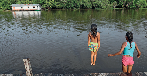 Imagem: Fotografia. Duas crianças vistas de costas. Uma dela está saltando para local com água de cor verde, ela tem cabelos longos pretos, sem camiseta e com bermuda verde com estampas coloridas e mãos sobre as coxas. À direita, uma menina vista de costas, de cabelos pretos grandes, vestida com blusa regata em azul-claro, short curto rosa, perto de local com madeira. Ao fundo, depois das águas, vegetação e uma casa de paredes brancas, com telhado cinza e três janelas.  Fim da imagem.