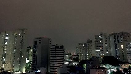 Imagem: Fotografia. A mesma cidade descrita anteriormente, com céu de cor cinza, prédios com luzes acesas.  Fim da imagem.