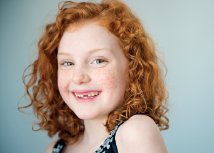 Imagem: Fotografia. Uma menina ruiva, de pele clara, vista dos ombros para cima. Ela tem cabelos encaracolados, para à esquerda, sorrindo, usando blusa de mangas curtas em preto. Fim da imagem.