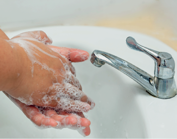 Imagem: Fotografia. Duas mãos de uma pessoa de pele morena, com bolhas de sabão esfregando, perto de uma pia branca com torneira de cor cinza.  Fim da imagem.
