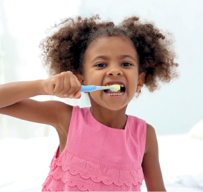Imagem: Fotografia. Uma menina de pele morena vista da cintura para cima. Ela tem cabelos crespos castanhos, usando blusa regata de cor rosa, olhando para frente escovando os dentes com escova na mão esquerda.  Fim da imagem.