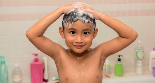 Imagem: Fotografia. Um menino lavando os cabelos com muitas espumas de cor branca. Ele é visto do busto para cima, de pele morena-claro, cabelos e olhos pretos. Ele está com os braços para cima, mãos nos cabelos. Ao fundo, produtos com embalagens coloridas.  Fim da imagem.