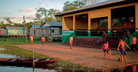 Imagem: Fotografia. Vista de local aberto com crianças jogando futebol sobre solo marrom, algumas delas estão vestidas com camiseta vermelha. À esquerda, contorno com grama de cor verde, uma canoa de madeira de cor marrom. Ao fundo, uma escadaria onde algumas crianças estão sentadas vendo o jogo. Em segundo plano, à esquerda, casas com os telhados triangulares e árvores de folhas verdes.  Fim da imagem.