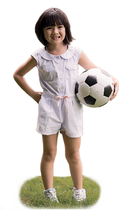 Imagem: Fotografia. Uma menina em pé em local com grama verde. Ela tem pele clara, cabelos curtos pretos com franja, com macacão branca de regata e short e sapatos brancos. Ela está com a mão esquerda na cintura e segura na mão direita uma bola de futebol em branco e preto, olha para frente sorrindo.  Fim da imagem.