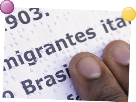 Imagem: Fotografia. Um texto em preto sobre base branca, com pequenos círculos em relevos, por onde passa dois dedos de pele morena e unhas curtas.  Fim da imagem.