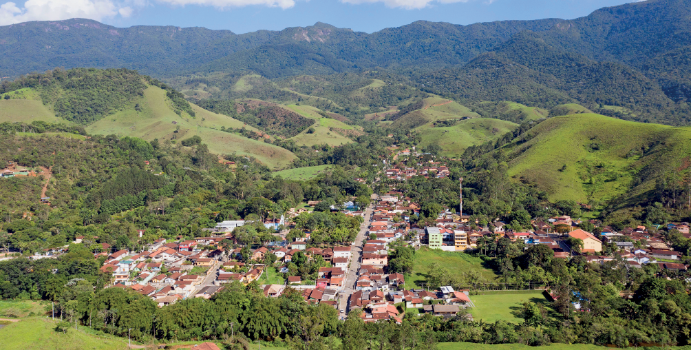 Imagem: Fotografia. Vista geral de local com cidade ao centro, composta casas próximas umas das outras, muitas com telhados de cor marrom, entre árvores com folhas verdes e ao fundo, morros cobertos com vegetação em verde-claro e partes em verde-escuro. Na parte superior, céu azul-claro com nuvens brancas.  Fim da imagem.