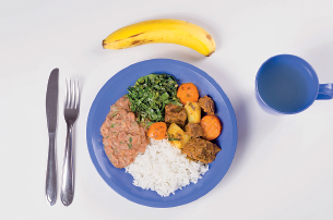 Imagem: Fotografia. Vista do alto, um prato redondo e copo de cor azul. No prato, comida : arroz, legumes e verdes, carne. À esquerda, faca e garfo cinza e na parte superior, banana.  Fim da imagem.