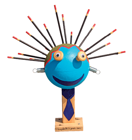 Imagem: Fotografia. Objeto com formato de rosto na vertical feito com vários elementos de cor marrom, com olhos em azul, nariz em azul-claro, boca em vermelho, com hastes finas nas extremidades na parte superior e bolinhas vermelhas na parte superior, um fio longo coloridos para esquerda e para à direita.  Fim da imagem.