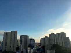 Imagem: Fotografia. Vista geral de cidade com vários prédios com sombra sobre eles e na parte superior, céu de cor azul-claro e com nuvens brancas à direita.  Fim da imagem.