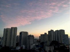 Imagem: Fotografia. Vista geral da mesma cidade todos os prédios com sombra sobre eles e na parte superior, céu de cor azul e nuvens em tons de rosa. Fim da imagem.