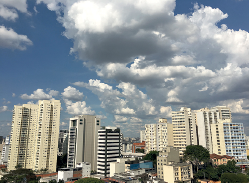 Imagem: Fotografia. Vista geral de cidade da mesma cidade com vários prédios de cor bege-claro e branco e luz sobre eles, com o céu em azul-claro e muitas nuvens brancas.  Fim da imagem.