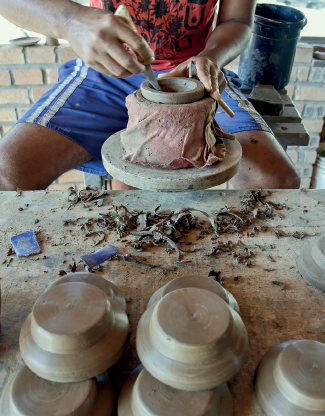 Imagem: Fotografia. Vista parcial de homem agachado, visto parcialmente, fabricando um vaso de cor marrom. À frente, com um utensílio na mão esquerda. Ele usa blusa de cor vermelha, bermuda de cor azul, com listas em branco. Embaixo, pedaços e rebarbas. Mais à frente, outros vasos de cerâmicas igual. Fim da imagem.