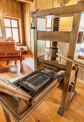 Imagem: Fotografia. Máquina de madeira na vertical, com uma engrenagem ao centro na vertical e à frente, hastes na horizontal. Mais ao fundo, local com paredes, janela de madeira. Fim da imagem.
