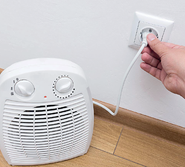 Imagem: Fotografia. Mão de uma pessoa de pele clara, colocando cabo branco de aparelho elétrico branco com botões em tomada na parede. O aparelho é pequeno, com formato na vertical. Na parte inferior, chão de madeira de cor bege.    Fim da imagem.