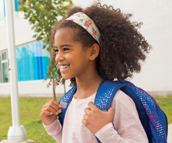 Imagem: Fotografia. Menina vista do busto para cima, virada para à esquerda, com pele negra, com cabelos crespos de cor marrom, com faixa de cor branca com estampa florida, usando blusa de mangas compridas em rosa-claro, com mochila de cor azul nas costas, com as mãos sobre as alças da mochila, olhando para frente sorrindo. Ao fundo, local com grama de cor verde, vegetação e parede branca com janelas.  Fim da imagem.
