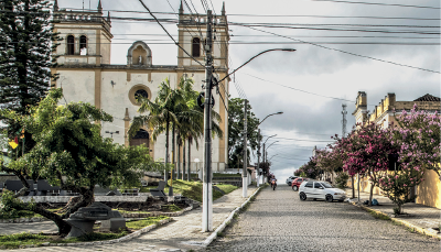 Imagem: Fotografia. Ao centro, rua de cor cinza, com calçada à esquerda e à direita e um carro passando na rua. À esquerda, vegetação verde com árvores com folhas longas. Na calçada, lâmpadas. Mais ao fundo, catedral de paredes de cor branca com detalhes de cor bege. Na parte superior, céu de cor cinza, nublado. Na parte superior, cabos de energia preta.  Fim da imagem.
