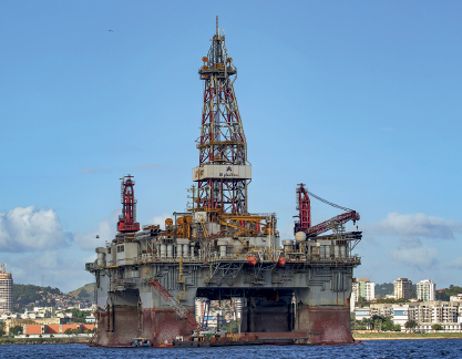 Imagem: Fotografia. Uma plataforma em alto mar, com base na horizontal na vertical e ao centro, uma torre central de ferro. À esquerda e à direita, partes de ferro. No mar, água de cor azul-escuro. Em segundo plano, cidade com prédios e morros. Na parte superior, céu de cor azul-claro com algumas nuvens brancas.  Fim da imagem.