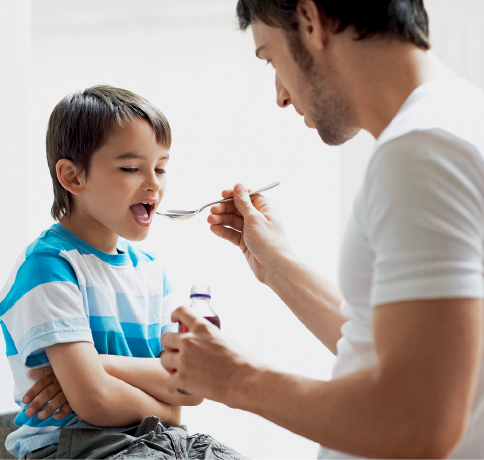 Imagem: Fotografia. À esquerda, um menino sentado de pele clara, cabelos castanhos lisos, usando camiseta com listras em azul e branco e calça jeans, com os braços cruzados. À frente, um homem dando remédio com colher na mão direita. Ele tem pele clara, cabelos pretos, com camiseta de mangas curtas.  Fim da imagem.