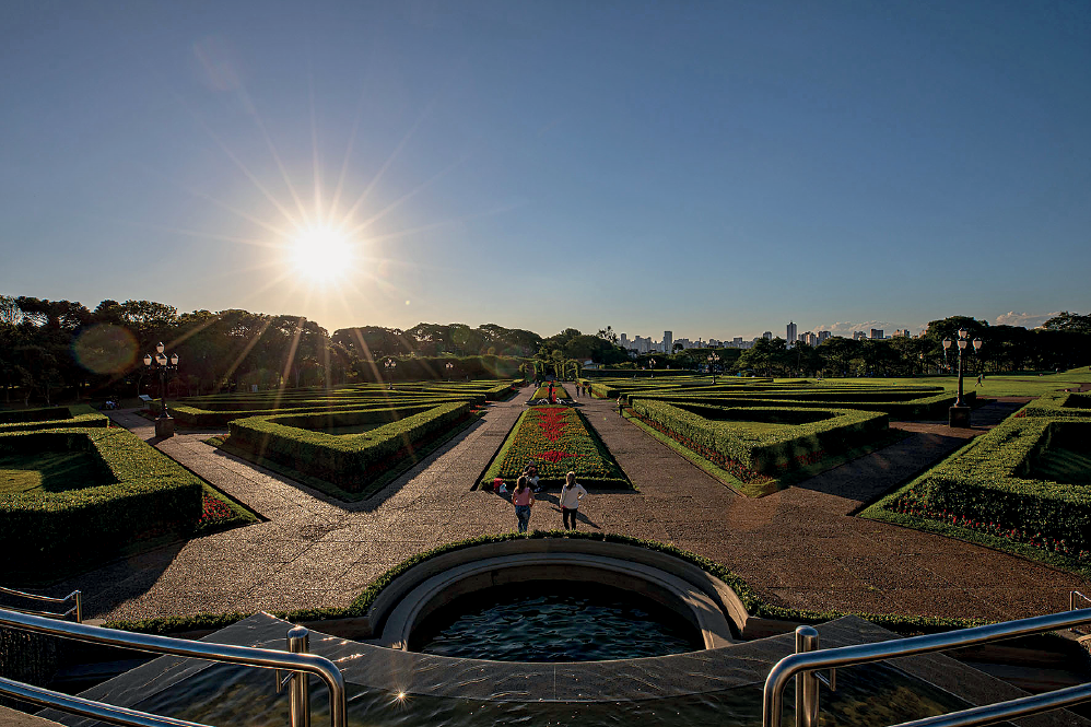 Imagem: Fotografia. Vista geral de local do alto. À direita, corrimão cinza na horizontal. Mais ao centro, uma fonte de água com parte quadrada e parte inferior arredondada. Mais ao fundo, local com alas grandes e jardim decorado com vegetação alta cortada em formato triangular e quadrado. Mais ao fundo, árvores de folhas verdes. Na parte superior, céu em azul-claro com sol à esquerda em amarelo incandescente.  Fim da imagem.