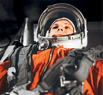 Imagem: Fotografia. Vista da cintura para cima de homem de pele clara, com capacete arredondado em branco, usando roupa de astronauta de cor laranja, com cintos cinzas sobre o corpo na vertical na diagonal.  Fim da imagem.