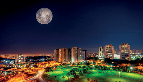Imagem: Fotografia. Vista geral de local, cidade com área arborizada à direita com árvores com folhas verdes. À esquerda, rodovias com luz de cor amarela e ao fundo, prédios altos com luzes acesas. Na parte superior, céu azul-escuro com uma lua grande à esquerda, de cor branca, com partes em cinza.  Fim da imagem.