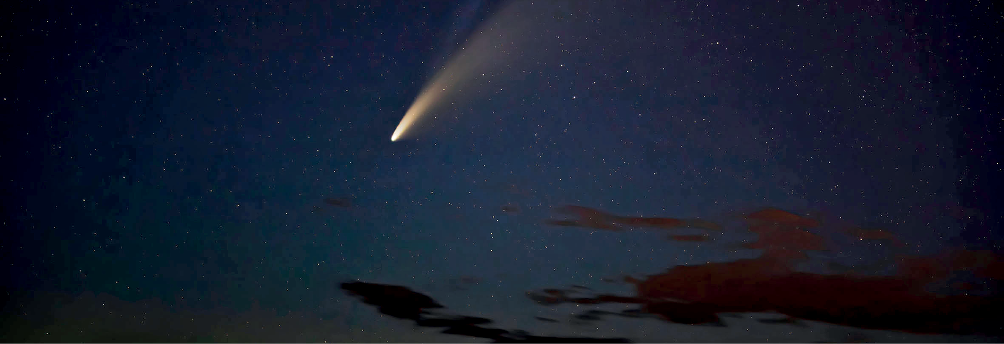 Imagem: Fotografia noturna. Céu de cor azul-escuro, com nuvens escuras na ponta inferior à direita, com pequenos pontos brancos coloridos. Na parte superior, cometa na vertical de cor branca com parte superior em bege.  Fim da imagem.