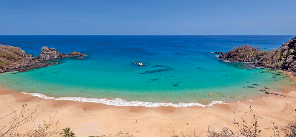 Imagem: Fotografia. Vista do alto de praia, com areia de cor bege-claro na parte inferior e mais ao fundo, oceano de cor verde-claro na borda e ao fundo, em azul. Na ponta da esquerda e à direita, pedras em marrom. Acima, céu de cor azul-claro.  Fim da imagem.