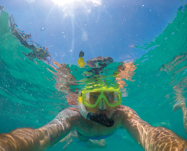 Imagem: Fotografia. Um homem de pele clara, dentro do mar de cor verde-claro, tirando selfie dele, com os braços esticados. Ele está com uma máscara de mergulho em amarelo com tubo na boca em preto e parte transparente na vertical.  Fim da imagem.