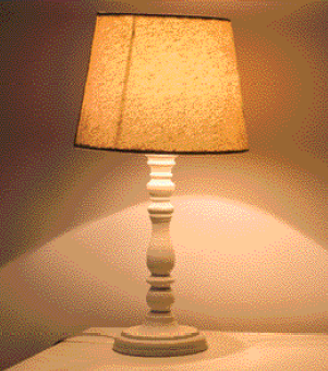 Imagem: Fotografia. Um abajur de cor bege na vertical e parte superior, forma arredondada de cor amarela onde é possível ver parte da luz acesa.  Fim da imagem.