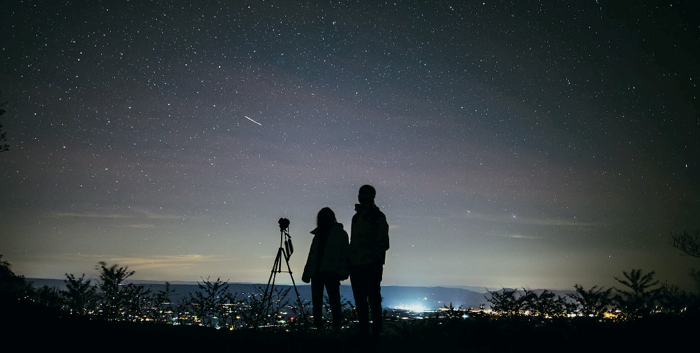 Imagem: Fotografia noturna. À esquerda, pedestal sobre telescópio, com duas pessoas à direita, sendo vistas como silhuetas, uma menor com casaco de mangas compridas e outra pessoa mais alta, com casaco com mangas compridas. Ao redor, luzes ao longe da cidade e árvores com folhas. Na parte superior, céu de cor azul-escuro com centena de pontos brilhantes em branco no céu.  Fim da imagem.