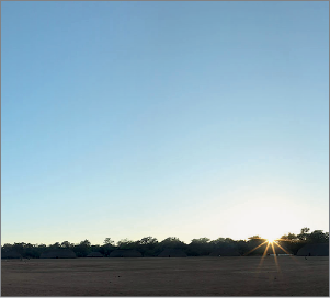 Imagem: Fotografia. O mesmo local descrito anteriormente, com sombra no solo, com sol visto de tamanho menor e menos forte, à direita. O céu é de cor azul-claro.  Fim da imagem.